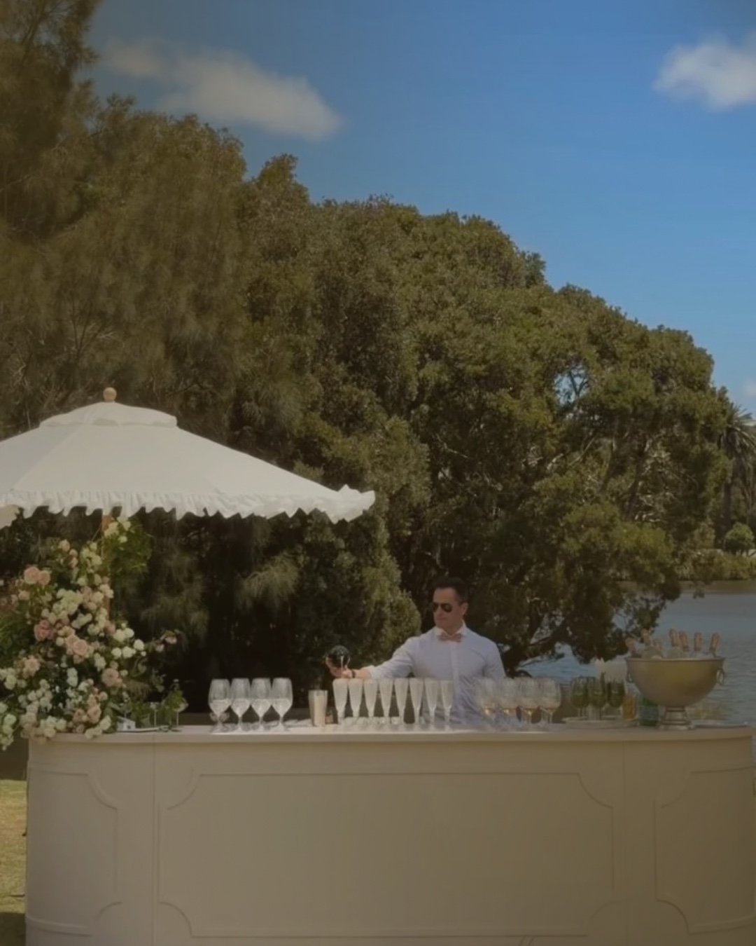 Barman derrière un bar mobile blanc, avec verrerie et fleurs, dans un jardin au bord de l’eau